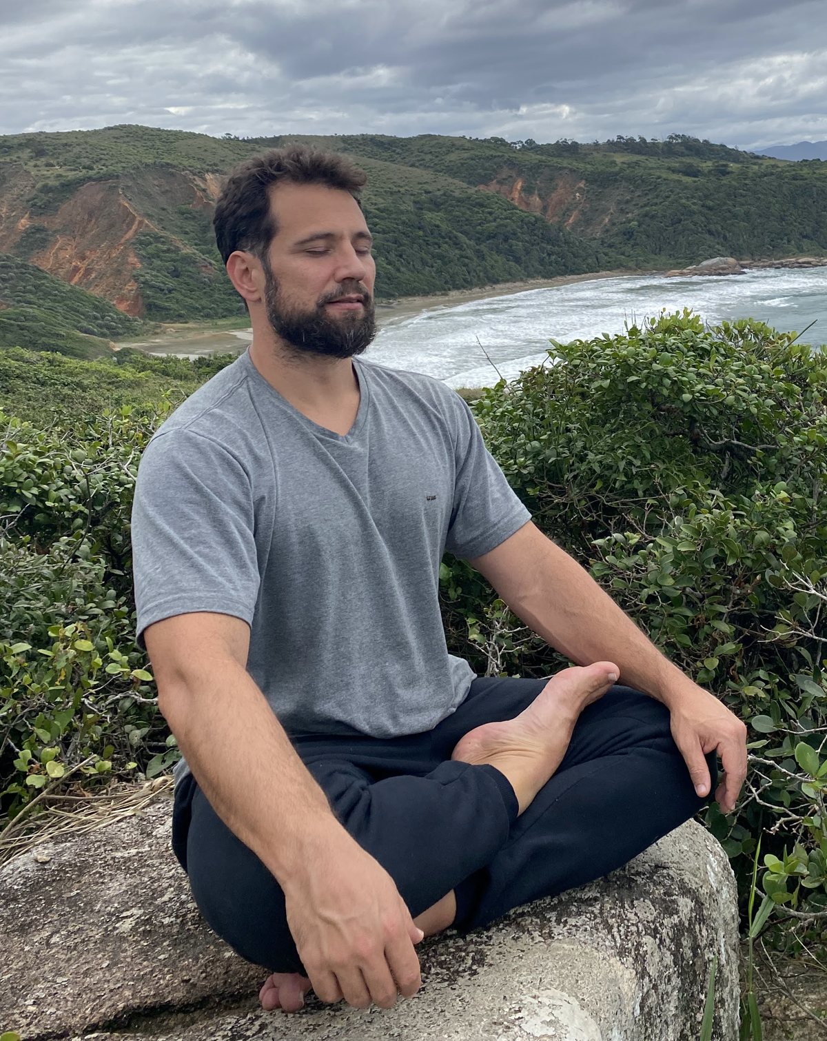 Prof. Daniel De Nardi em medita&ccedil;&atilde;o sobre uma pedra, com a praia ao fundo